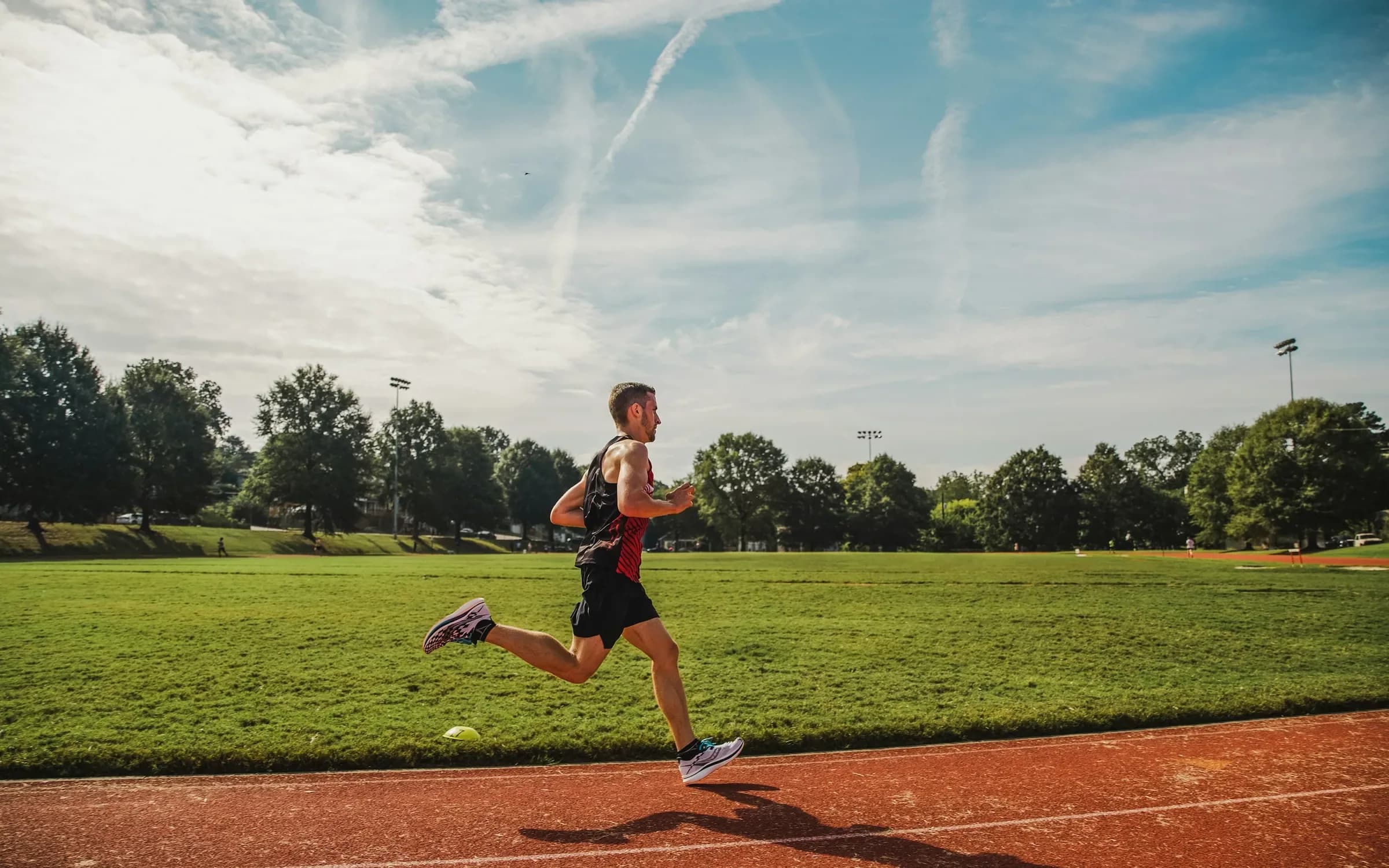 A man running on a red track beside a green field under blue sky